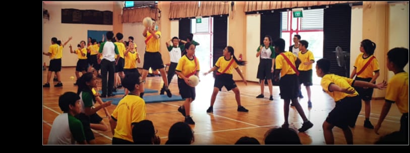 Students in yellow jerseys play a ball game in an indoor gymnasium with wooden floors.